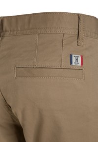 Primer plano de un bolsillo trasero de pantalones beiges con un lazo para cinturón y una pequeña etiqueta rectangular de tela tricolor que dice "Hilfiger H Nueva York".