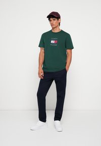 Grøn bomulds t-shirt med et trykt logo og navy bukser. Hvide sneakers fuldender outfittet sammen med en mørk maroon cap.