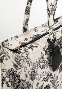 Sac en toile avec un fond beige et un motif floral noir, doté d'une fermeture à glissière sur le dessus, de deux poignées et d'une étiquette en cuir noir.