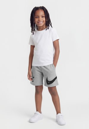 Barn med flettet hår, smilende, står med den ene hånd i lommen, iført hvid t-shirt, grå shorts med sort logo og hvide sneakers.