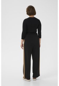 Sieviete stāv ar muguru pret kameru, valkājot melnu garroku tērpu un melnas platām kāju biksēm ar bēšām sānu svītrām un bēšām sporta apaviem.