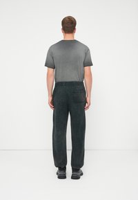 Mann, der mit dem Rücken zur Kamera steht und ein graues Kurzarmhemd, schwarze elastische Hose mit Gesäßtasche und dunkle Sneakers auf einem weißen Hintergrund trägt.