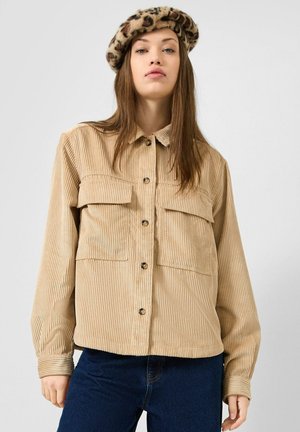 Femme portant une chemise beige en velours côtelé boutonnée et un béret à imprimé léopard, debout devant un fond clair uni.