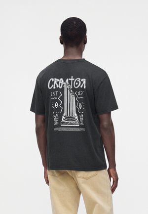 Mand iført en sort t-shirt med en hvid grafik af en brækket søjle og ordet "Creator" på bagsiden, kombineret med beige bukser.