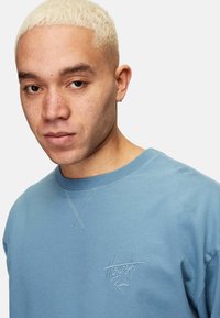 Ljusblå sweatshirt tillverkad av mjukt tyg. Har rund halsringning och broderad "Ärlighet Råder"-logotyp på vänster bröst. Enkel design.