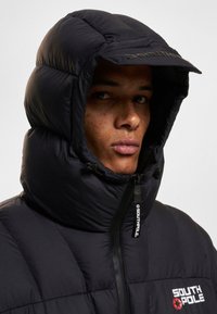 Veste en duvet noire avec un col haut et une grande capuche rembourrée. Présente une fermeture éclair à l'avant et le logo 'SOUTHPOL' en blanc sur la poitrine.