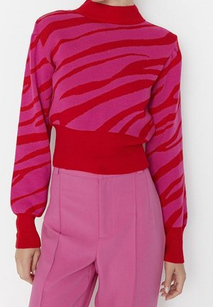 Mujer vistiendo un suéter de manga larga con un patrón rojo y rosa, metido dentro de pantalones de cintura alta color rosa, sobre un fondo blanco.