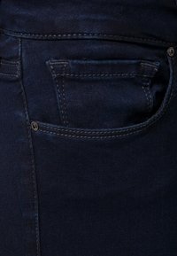 Los jeans de denim azul oscuro presentan un pequeño bolsillo trasero con costuras visibles y un acento de botón metálico. La tela tiene una textura suave.