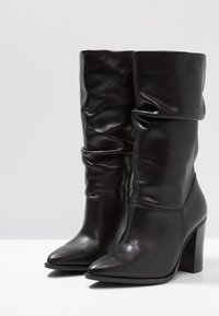 Botas de cuero negro hasta la rodilla con un diseño despeinado, puntera afilada y tacón alto apilado. Textura suave con detalles de costura mínimos.
