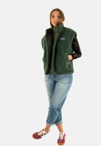 Gilet in pile verde con colletto alto, chiusura a bottoni e tasche laterali, abbinato a jeans azzurri e sneakers bordeaux.