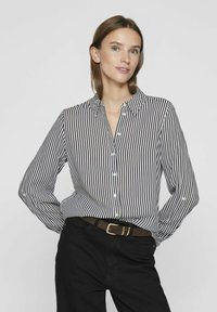 Woman wearing black pants and a black-and-white vertical striped button-up shirt, standing with hands behind back against plain background.