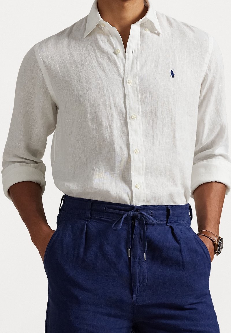 Mężczyzna w białej lnianej koszuli zapinanej na guziki z podwiniętymi rękawami, granatowych spodniach ze ściągaczem i zegarkiem na lewym nadgarstku.