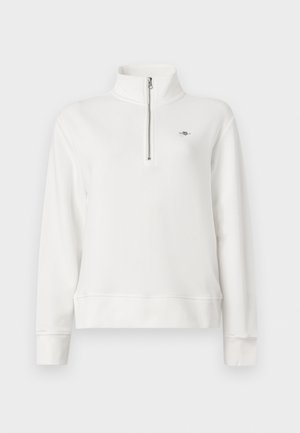 Bijeli pulover s dugim rukavima, prednjim rajsferšlusom do četvrtine i visokim ovratnikom, s malim izvezenim logotipom na lijevoj strani prsa.