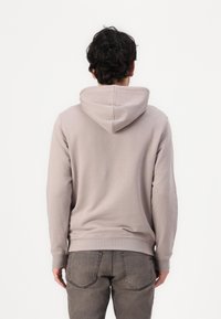 Ανοιχτό καμηλό φούτερ με κουκούλα και κορδόνι, με ριμπε διακοσμητικά στα μανίκια και στο κάτω μέρος. Φορεμένο με γκρι τζιν, παρουσιάζοντας έναν απλό, διακριτικό σχεδιασμό.