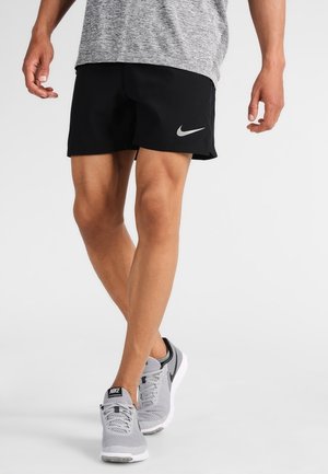 Persoon draagt zwarte Nike hardloopshorts, grijze Nike hardloopschoenen en een grijs sportshirt, staand met gekruiste benen tegen een effen achtergrond.