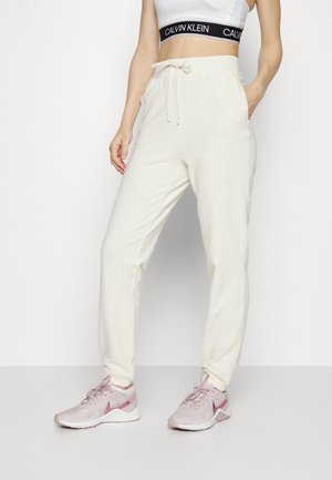 Femme portant un jogging blanc à cordon, un crop top blanc avec une bande noire Calvin Klein et des baskets de sport roses, debout avec une main dans la poche.