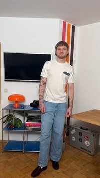 Mladý muž s tetováním, v bílém tričku a džínech, stojící v moderním pokoji s televizí, oranžovou lampou, fotoaparátem a barevnými pruhy na zdi.