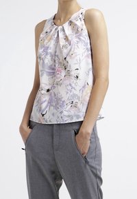 Ärmlös blommig blus i pastellfärger med plisserad halsringning, bärs med grå byxor som har dragkedjefickor. Slät tygtextur.