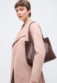 Καφέ υφαντή δερμάτινη τσάντα ώμου με διπλές λαβές, συνοδευόμενη από ένα απαλό ροζ παλτό. Η τσάντα έχει δομημένο σχήμα και υφή επιφάνειας.