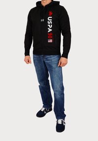 Felpa nera con zip e tasche frontali, con scritta e logo "USPA 90" in bianco e rosso. Indossata con jeans blu e sneakers scure.