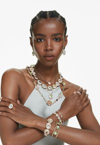 Goldfarbene Schmuckstücke, die mehrere bunte Edelsteine in verschiedenen Formen zeigen. Dazu gehören geschichtete Halsketten, Armbänder und Ringe mit filigranen Designs.