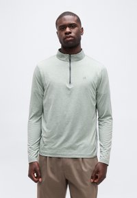 Άνδρας που φοράει ανοιχτό πράσινο μακρυμάνικο ρούχο με φερμουάρ και μπεζ παντελόνι, στέκεται μπροστά σε απλό λευκό φόντο.