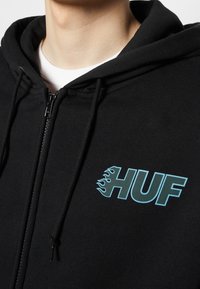 Felpa nera con zip, cappuccio regolabile e dettagli in fiamma blu sul logo "HUF", realizzata in tessuto morbido e texturizzato.