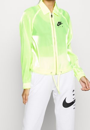 Osoba nosząca neonowo żółtą kurtkę Nike Air zapinaną na zamek błyskawiczny z białymi sznurkami oraz białe spodnie z wieloma czarnymi logo Nike.