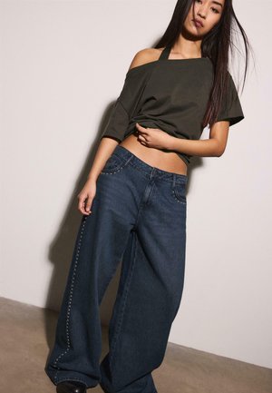 Jeune femme aux longs cheveux portant un haut foncé décolleté et un jean large en denim foncé orné de clous, debout contre un mur uni.