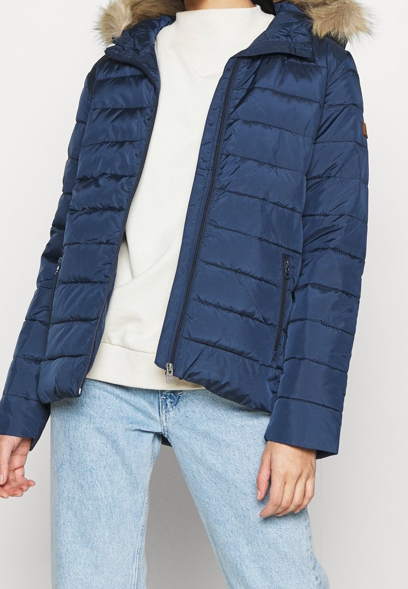 Personne portant une veste matelassée bleu marine avec une capuche bordée de fourrure, un pull blanc et un jean bleu clair, debout devant un fond uni.