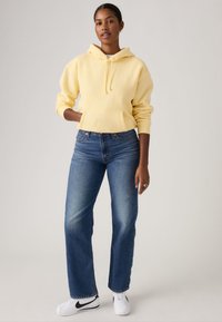 Κίτρινο φούτερ από βαμβάκι με μπροστινή τσέπη και κορδόνι, σε συνδυασμό με μπλε τζιν παντελόνι ίσιας γραμμής και λευκά αθλητικά παπούτσια με μαύρες λεπτομέρειες.