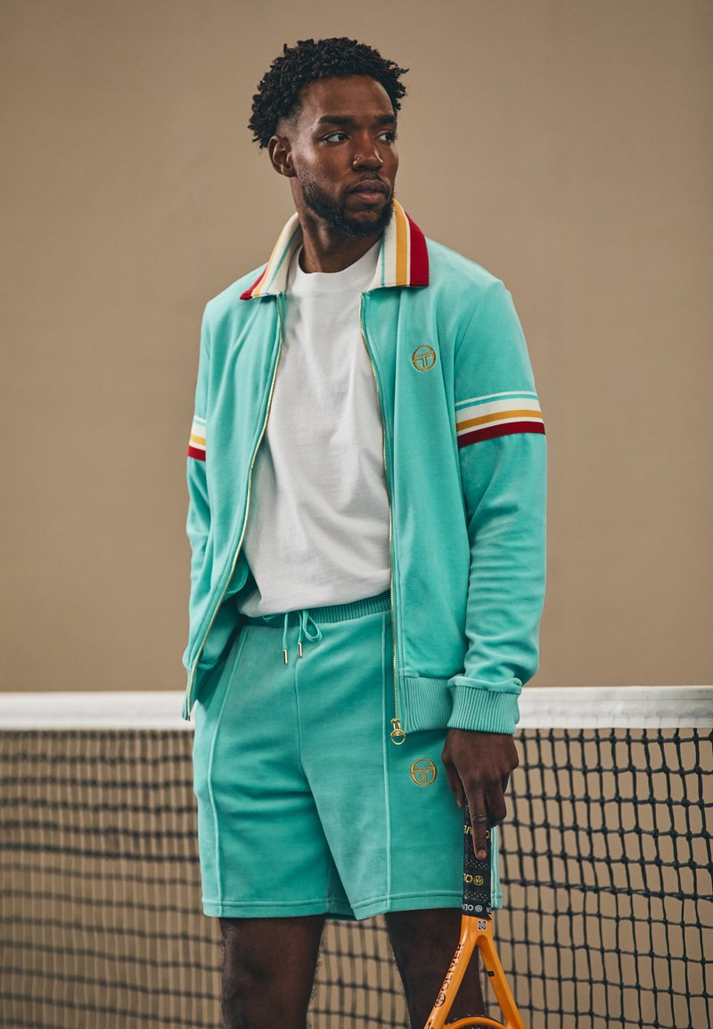 Veste de survêtement en velours teal avec des rayures rouges et jaunes, associée à un short assorti. Les accessoires comprennent un t-shirt blanc et une raquette de tennis.