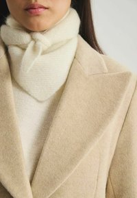 Manteau en laine beige au design structuré et à revers crantés, superposé sur un pull en laine blanc à col roulé et une écharpe crème nouée autour du cou.