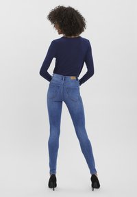 Jeans ajustados de mezclilla en azul, con una cintura alta, dos bolsillos traseros y una etiqueta beige en la cinturilla. Combinados con una blusa de manga larga en color marino.