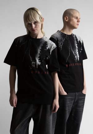 Dwie osoby noszące czarne koszulki z grafikami z białymi wzorami błyskawic i drzew oraz czerwonym tekstem, stojące na tle jednolitego jasnego tła.