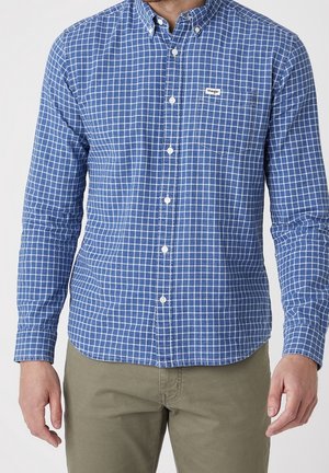 Mann trägt blau-weiß kariertes, langärmeliges Hemd mit Brusttasche und olivgrüne Hose vor einfarbigem Hintergrund.