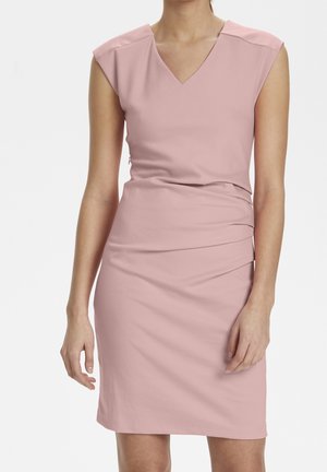 Femme portant une robe rose clair sans manches, longueur genou, avec un col en V et des détails drapés sur les côtés, debout devant un fond blanc uni.