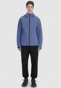 Personne debout face à l'avant, portant une veste bleue à capuche, un pantalon noir et des baskets noires sur un fond uni clair.