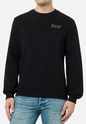Mann trägt schwarzen Langarm-Sweatshirt mit kleinem goldenem „Boxeur des Rues“-Logo auf der Brust und hellblaue Jeans.