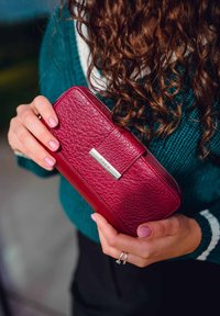 Red leather wallet with textured finish, silver hardware detail, and a zip closure. Held by a person wearing a green knitted sweater.