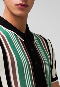 Pruhovaná polo košile s zelenými, hnědými, černými a bílými pruhy, s límečkem a knoflíkovou légou. Hladká pletená textura.