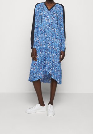 Osoba w niebieskiej, kwiecistej sukience midi z czarnymi długimi rękawami i białymi trampkami, stojąca na tle gładkiego jasnego tła.