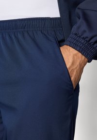 Pantaloni atletici navy cu o textură netedă, talie elastică și buzunare laterale. Materialul pare ușor și respirabil.