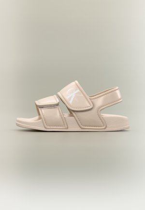 Μπεζ σανδάλι ανοιχτού δακτύλου με δύο ρυθμιζόμενα λουριά και ένα πίσω λουρί, με το λευκό λογότυπο "CK" της Calvin Klein στο επάνω λουρί.