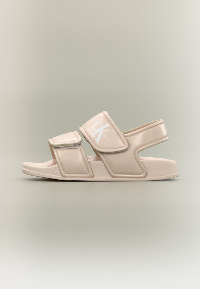 Μπεζ σανδάλι ανοιχτού δακτύλου με δύο ρυθμιζόμενα λουριά και ένα πίσω λουρί, με το λευκό λογότυπο "CK" της Calvin Klein στο επάνω λουρί.