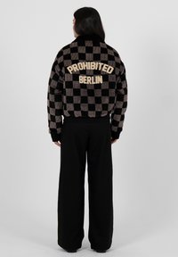 Person trägt eine schwarz-grau karierte Jacke mit dem Schriftzug "PROHIBITED BERLIN" auf dem Rücken und schwarze weite Hosen, steht mit dem Rücken zur Kamera.