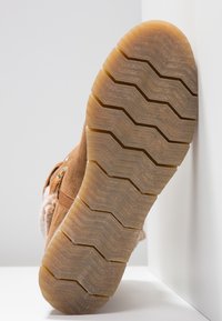 Semelle en caoutchouc avec motif en zigzag sur une botte d'hiver en daim beige avec doublure en fourrure, inclinée contre un mur blanc et un sol blanc.