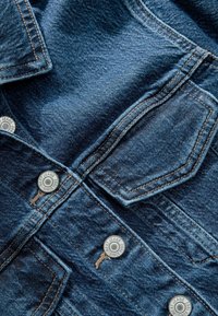 Gros plan sur un tissu en denim bleu avec des coutures détaillées, des boutons en métal portant l'inscription "Next Jeans Since 1982," et des rabats de poche visibles.
