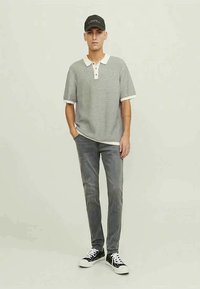 Camisa polo às riscas cinza e branca com colarinho bege, combinada com jeans cinza e ténis pretos. O modelo usa um boné preto.