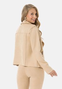 Veste en faux suède beige avec une coupe ajustée, des manches longues, un col cranté et des détails de couture au dos. Assortie d'un bas coordonné.